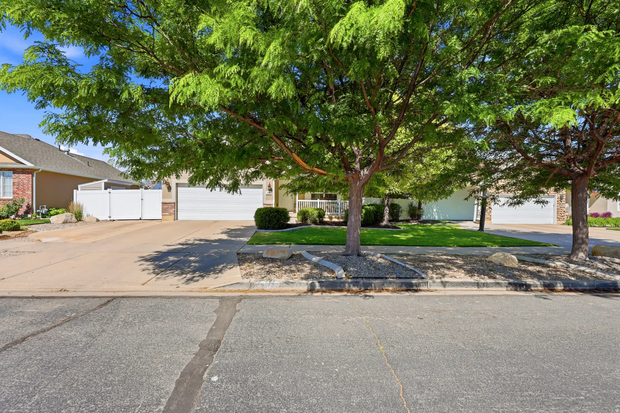 Manicured front yard with mature trees, a concrete driveway, and a white fence providing privacy