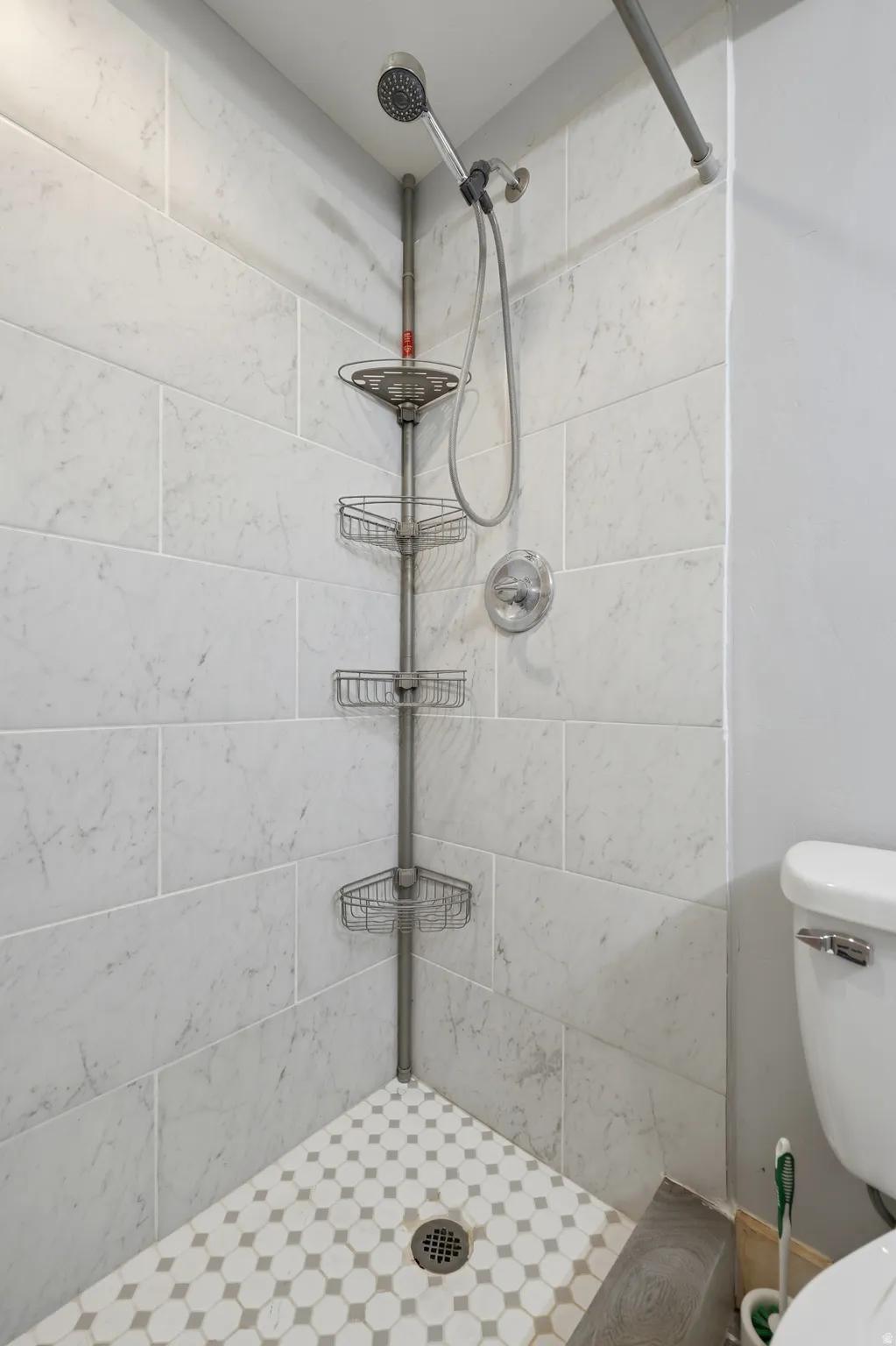 Tiled shower with a handheld showerhead, chrome fixtures, white subway tile walls, and a white and grey patterned mosaic tile floor