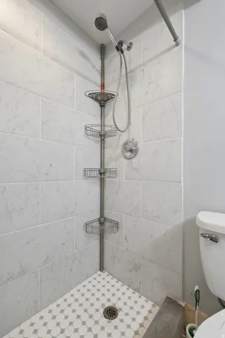 Tiled shower with a handheld showerhead, chrome fixtures, white subway tile walls, and a white and grey patterned mosaic tile floor