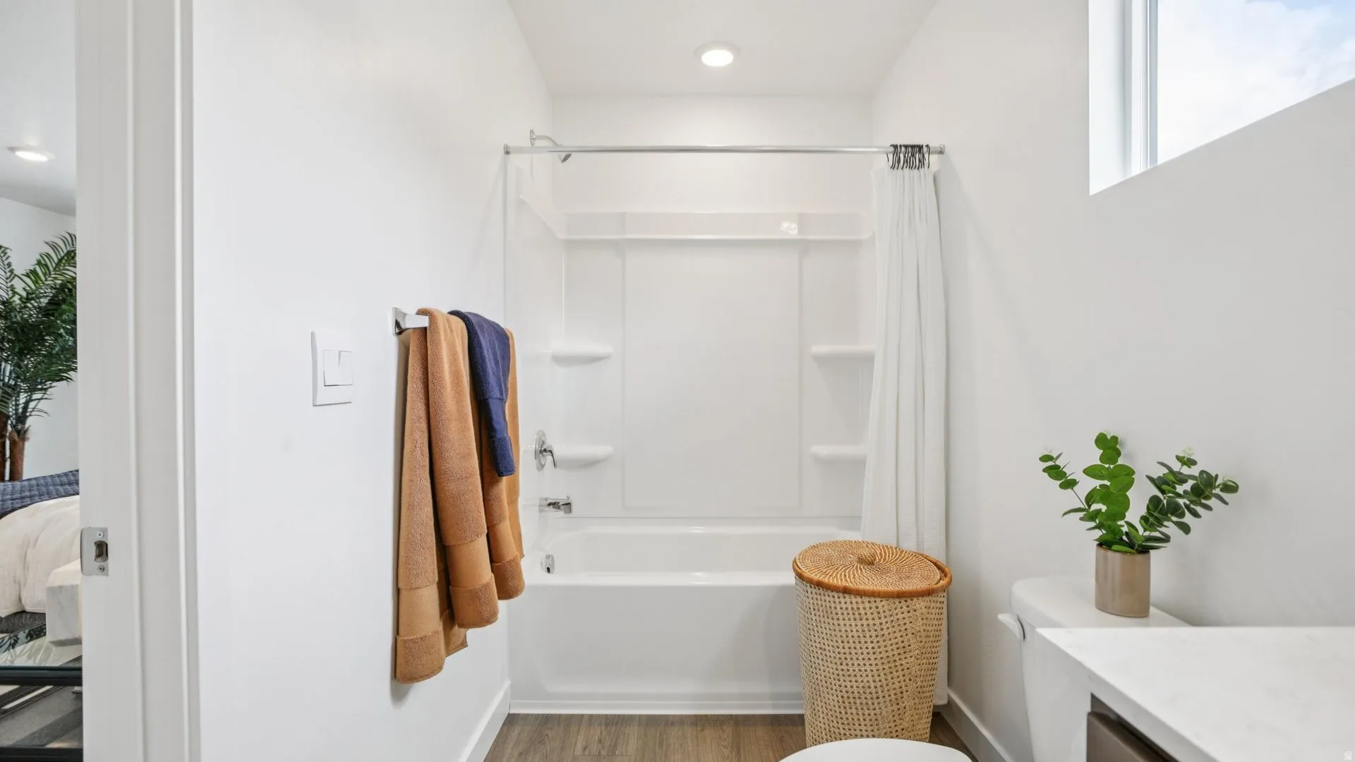 White bathroom featuring a built-in tub and shower unit, wood-finish flooring, a contemporary toilet, and a window providing natural light