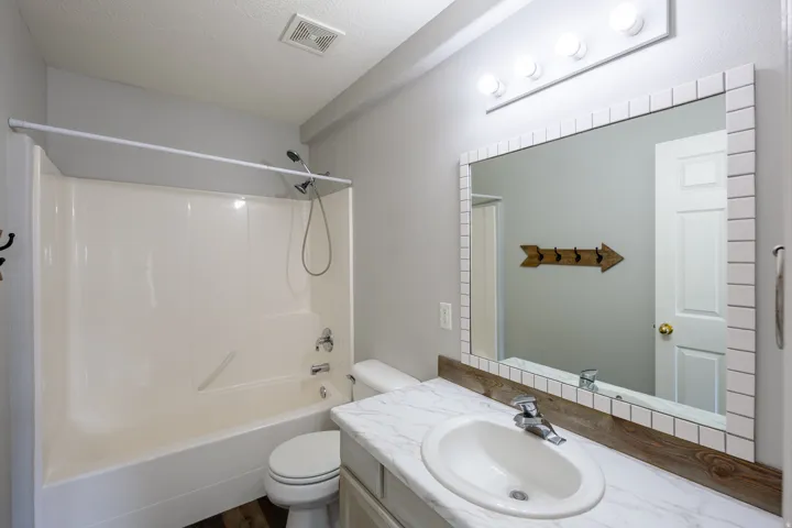 Bathroom featuring a white bathtub/shower combination, a vanity with a white countertop and integrated sink, a tiled mirror with a decorative frame, and overhead vanity lighting