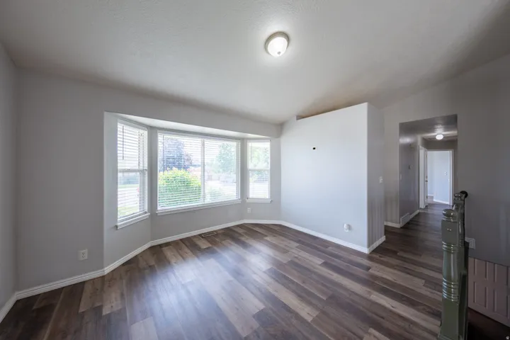 Spacious room featuring a bay window with blinds, wood-finish flooring, light gray wall paint, white baseboards, and a flush-mount ceiling light fixture