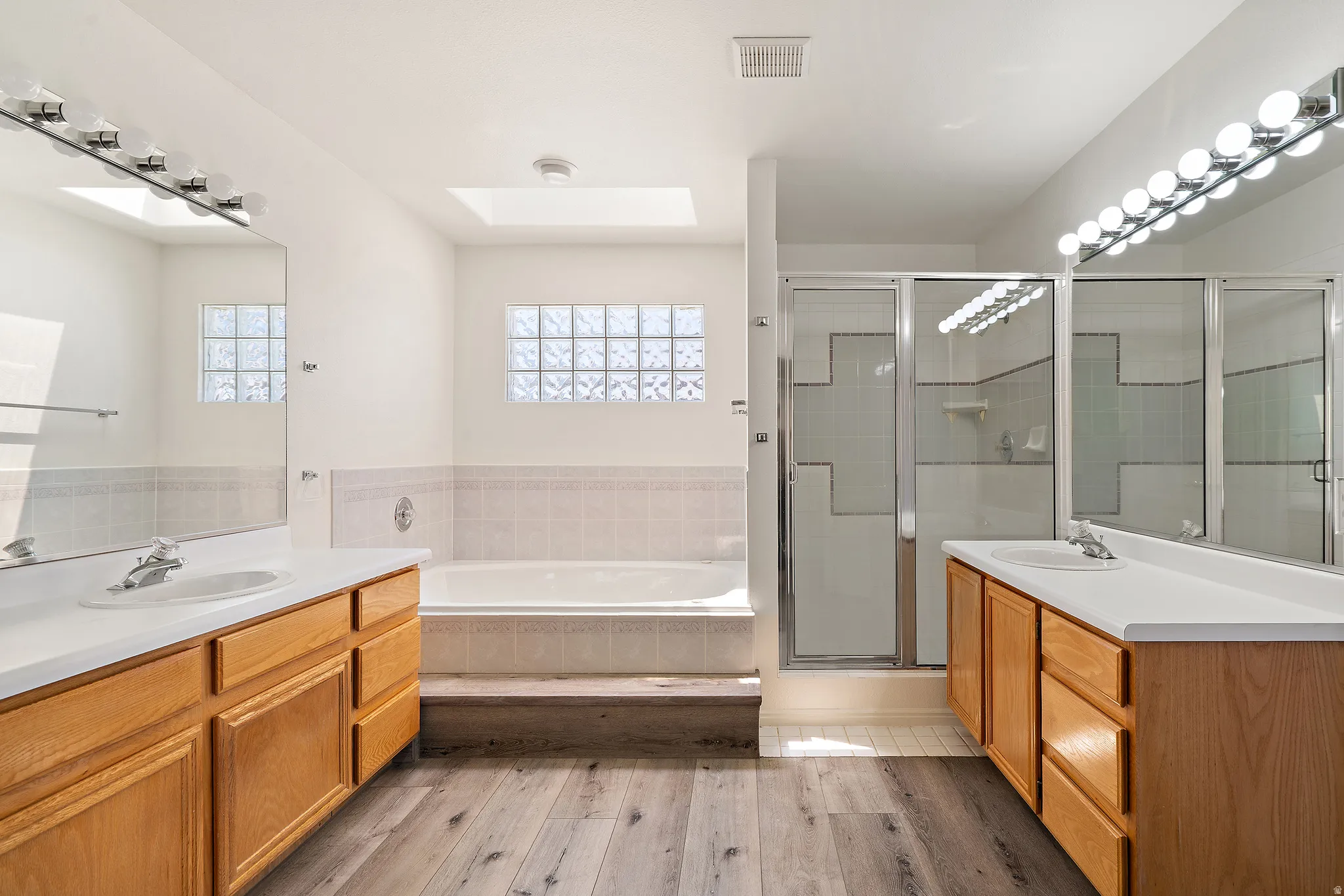 Bathroom featuring dual wood-finish vanities with integrated sinks, a built-in bathtub with tiled surround, a glass-enclosed shower, and wood-finish flooring