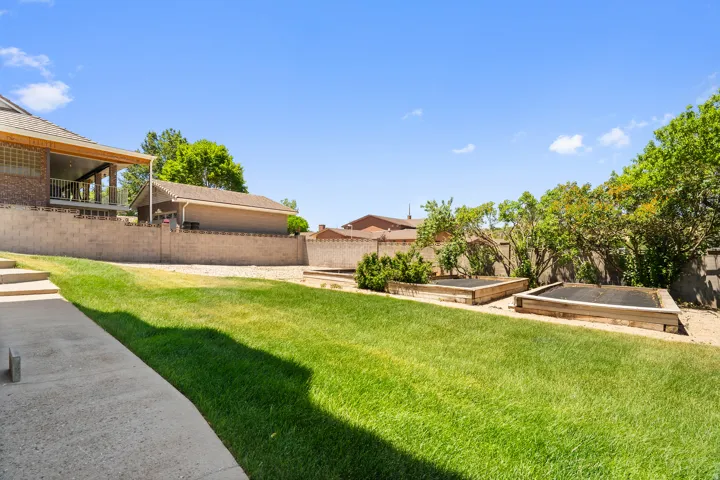 Expansive lawn with concrete pathway, raised garden beds, mature landscaping, and a covered patio structure