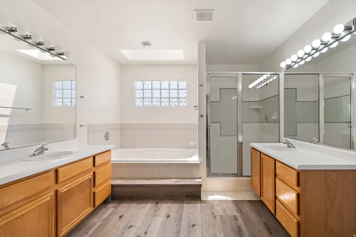 Bathroom featuring dual wood-finish vanities with integrated sinks, a built-in bathtub with tiled surround, a glass-enclosed shower, and wood-finish flooring