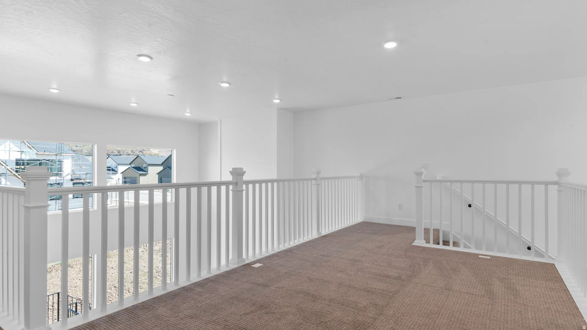 Spacious loft area featuring neutral carpeting, white wall finishes, recessed lighting, white balustrade railings, and multiple windows