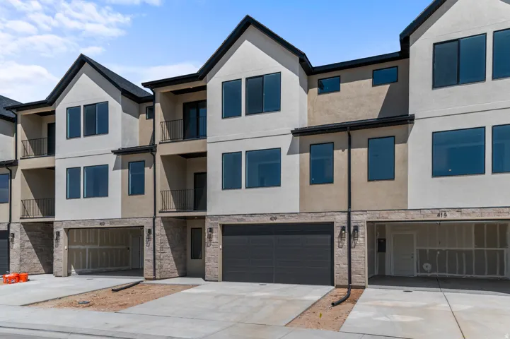 Contemporary townhome exterior featuring a neutral stucco and stone facade, black-framed windows, dark garage door, and a private balcony with black metal railings