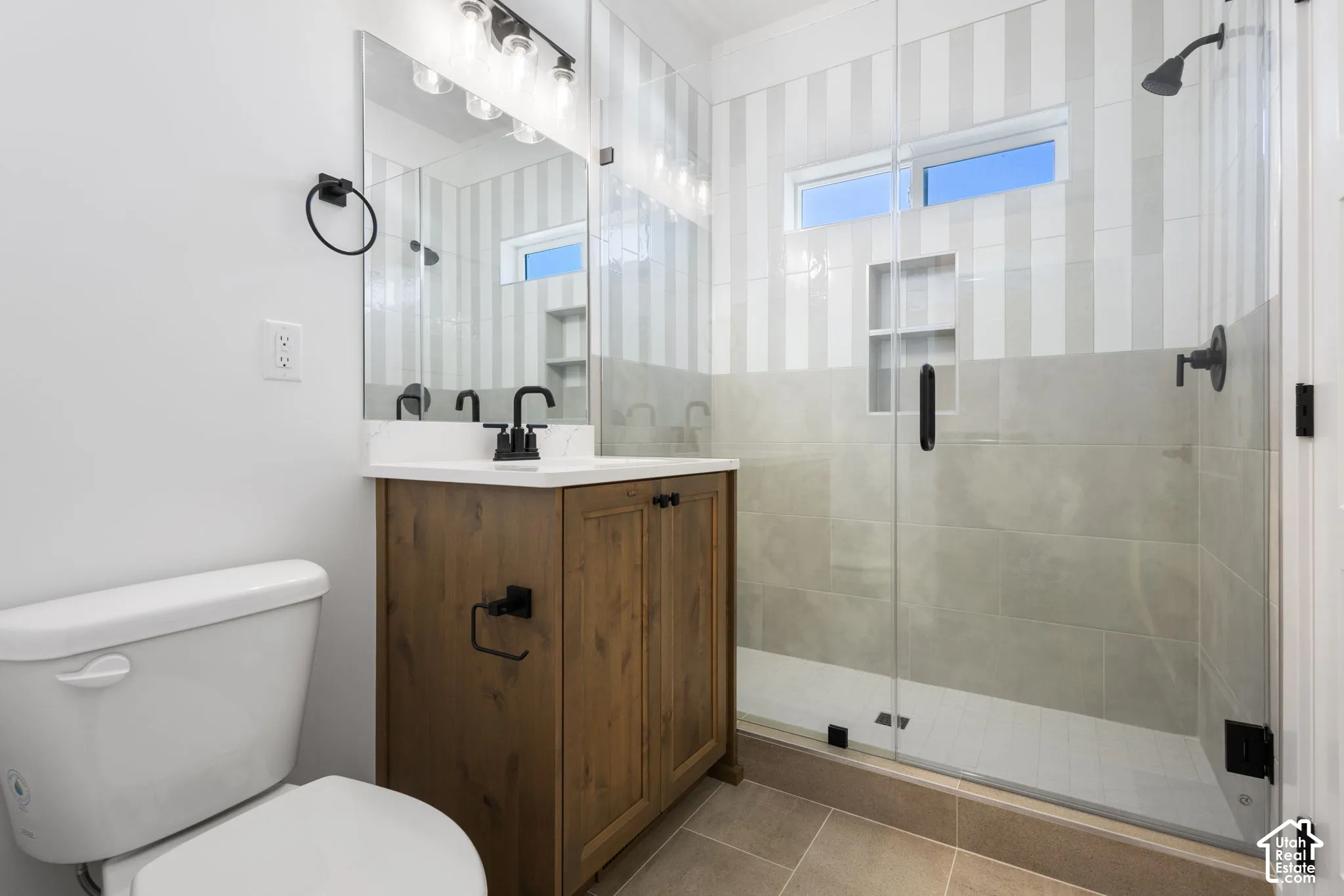Primary bedroom en-suite bathroom. Large glass-enclosed shower with a built-in niche, wood-finish vanity with a white countertop, and contemporary black fixtures.