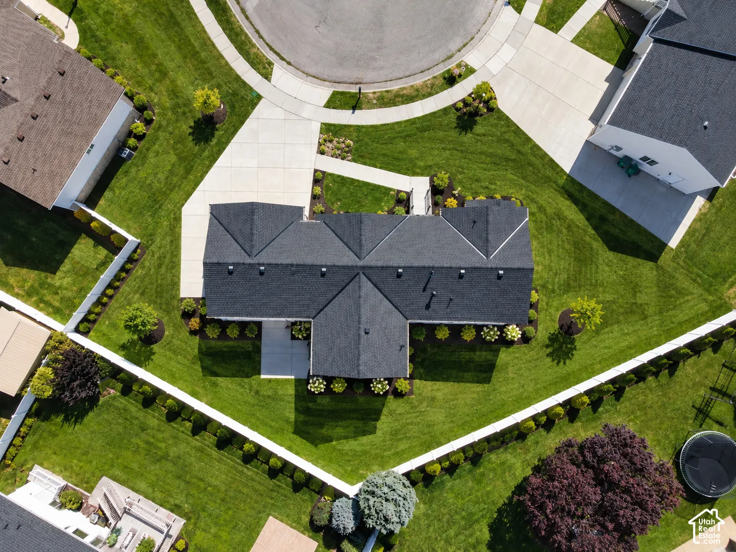 Manicured lawn with mature landscaping, extensive concrete driveway, dark shingle roofing, and a privacy fence