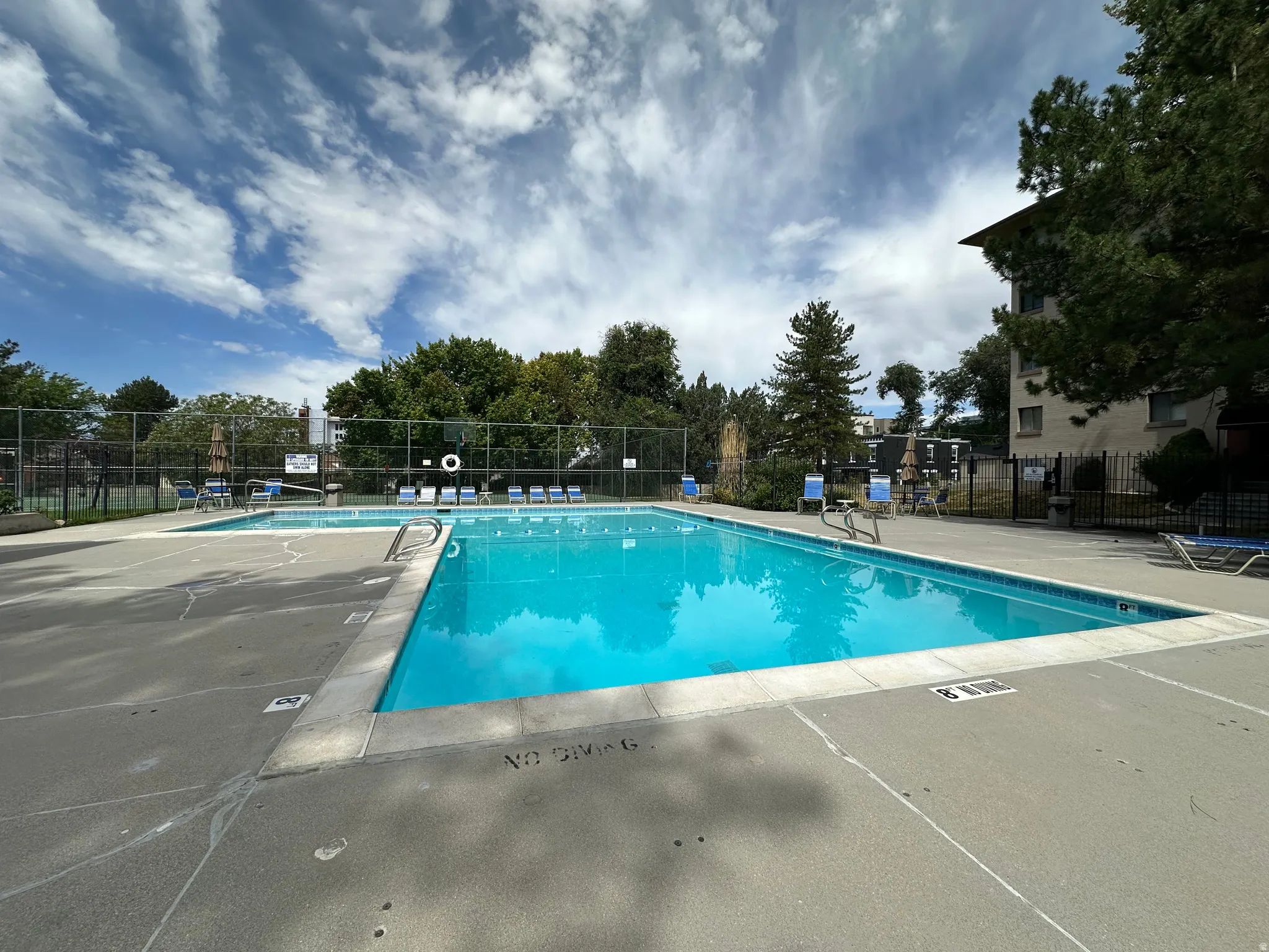 Outdoor swimming pool featuring a concrete deck surround, perimeter fencing, and mature trees