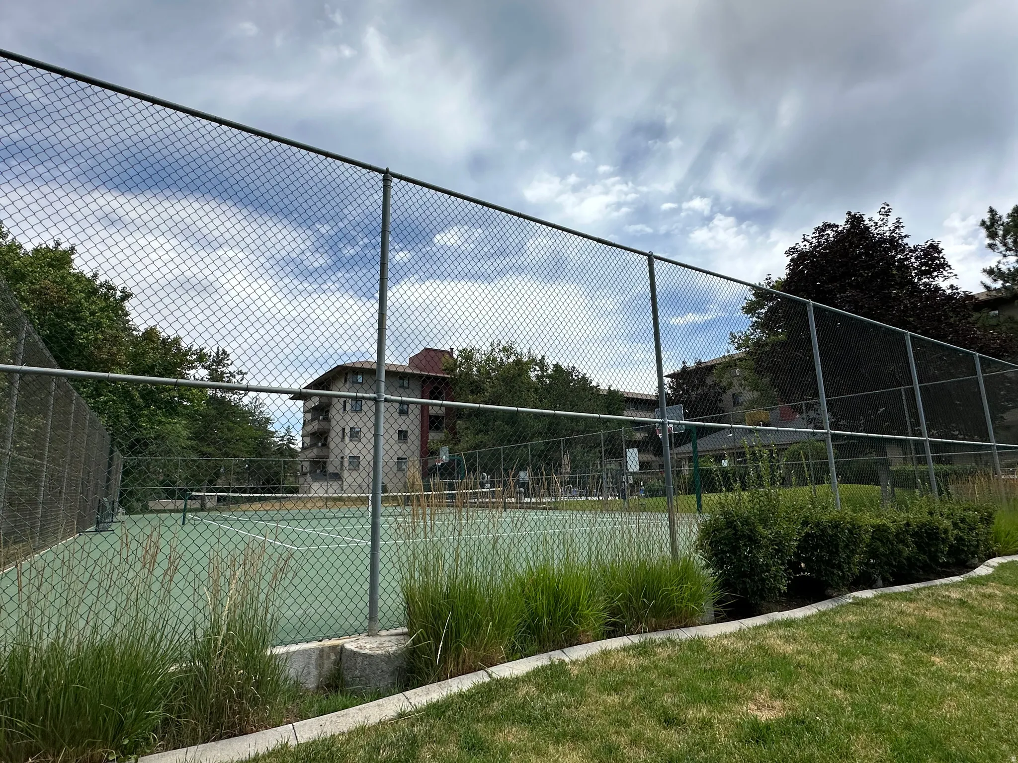 Outdoor tennis court with chain-link fencing, surrounded by mature trees and established landscaping