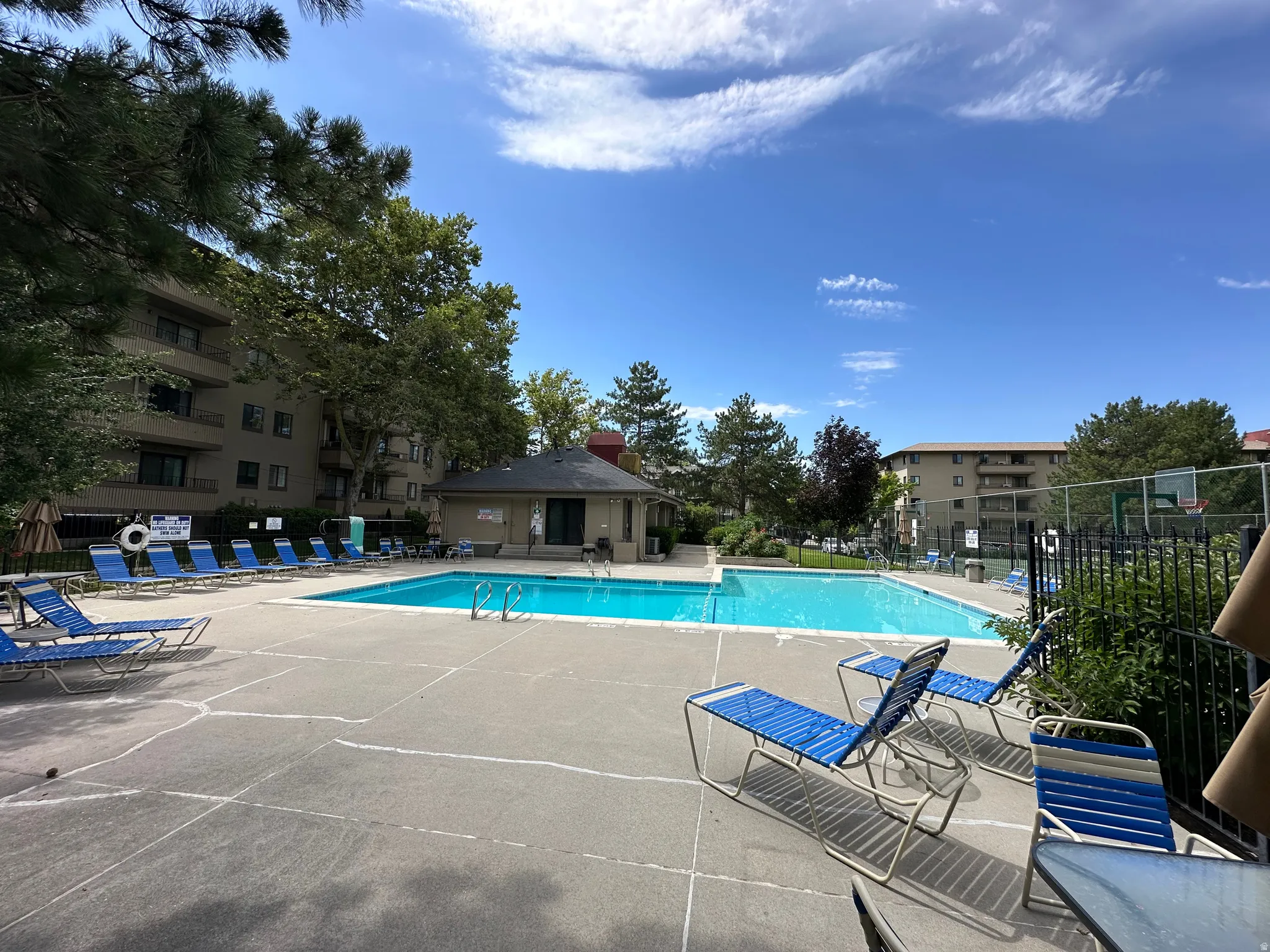 Outdoor swimming pool with surrounding concrete patio, adjacent pool house, and basketball court fencing
