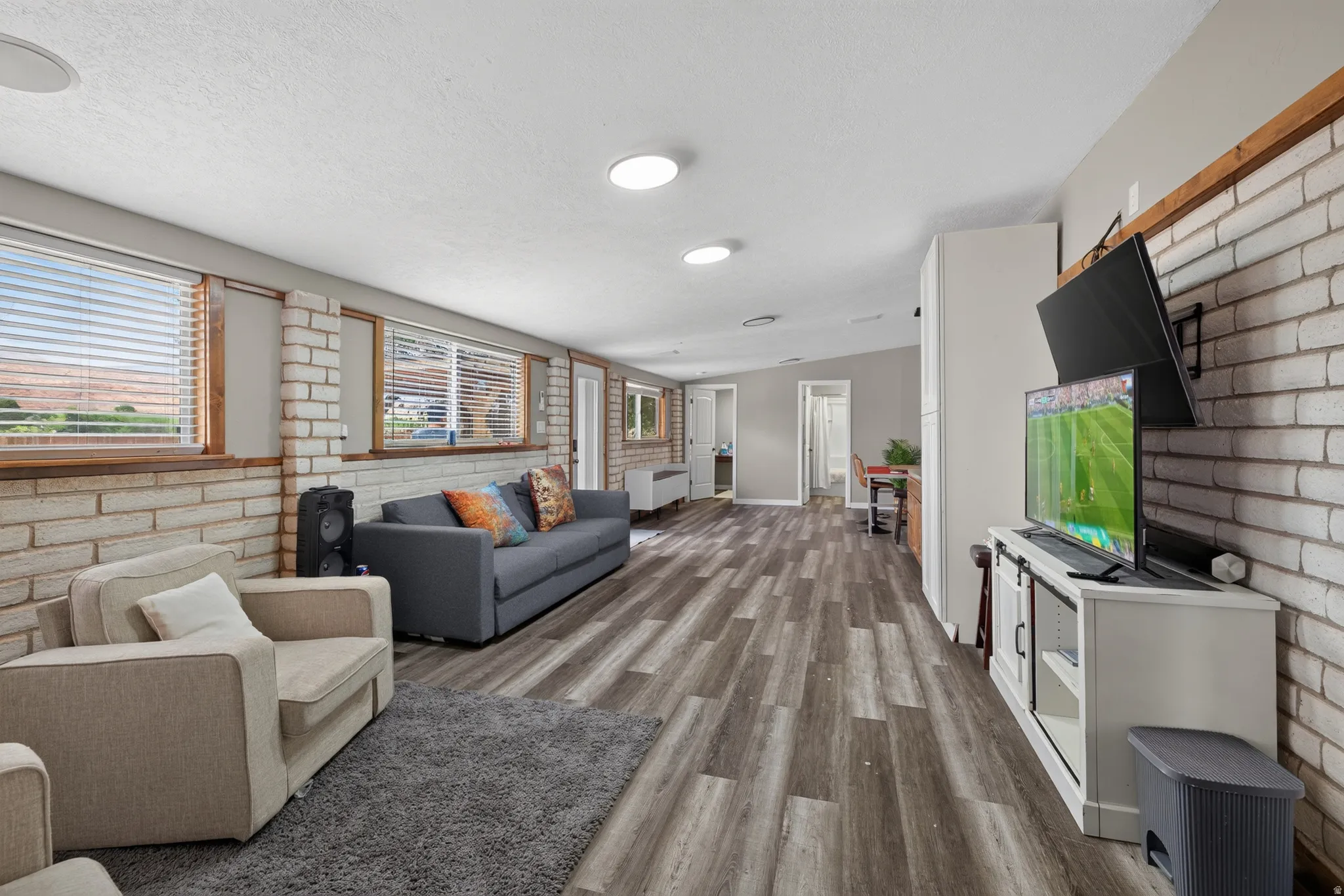 Spacious living area featuring wood-finish flooring, white brick-style walls, recessed lighting, and multiple windows with blinds