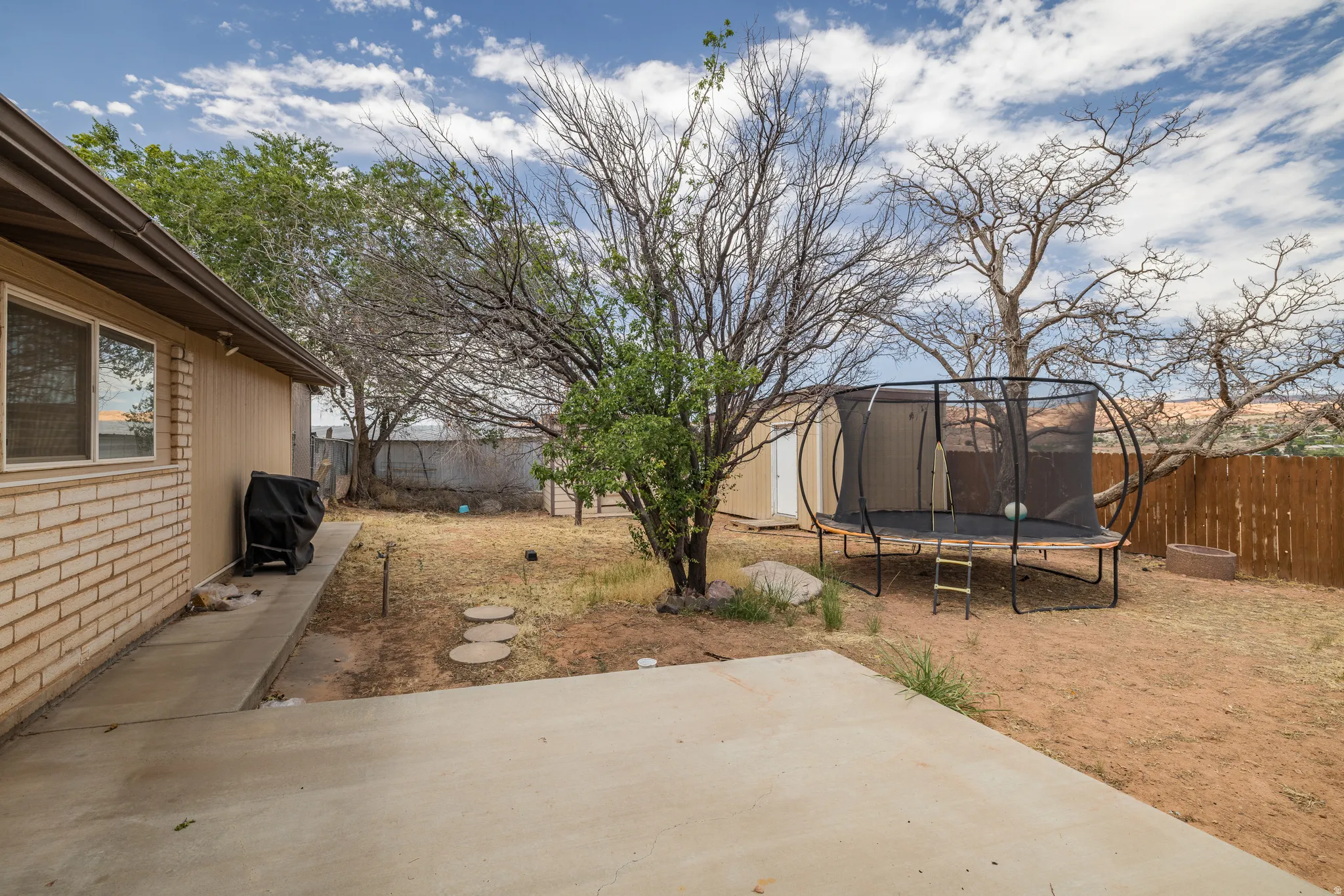 Spacious backyard with a concrete patio and walkway, featuring a mix of natural landscaping and mature trees