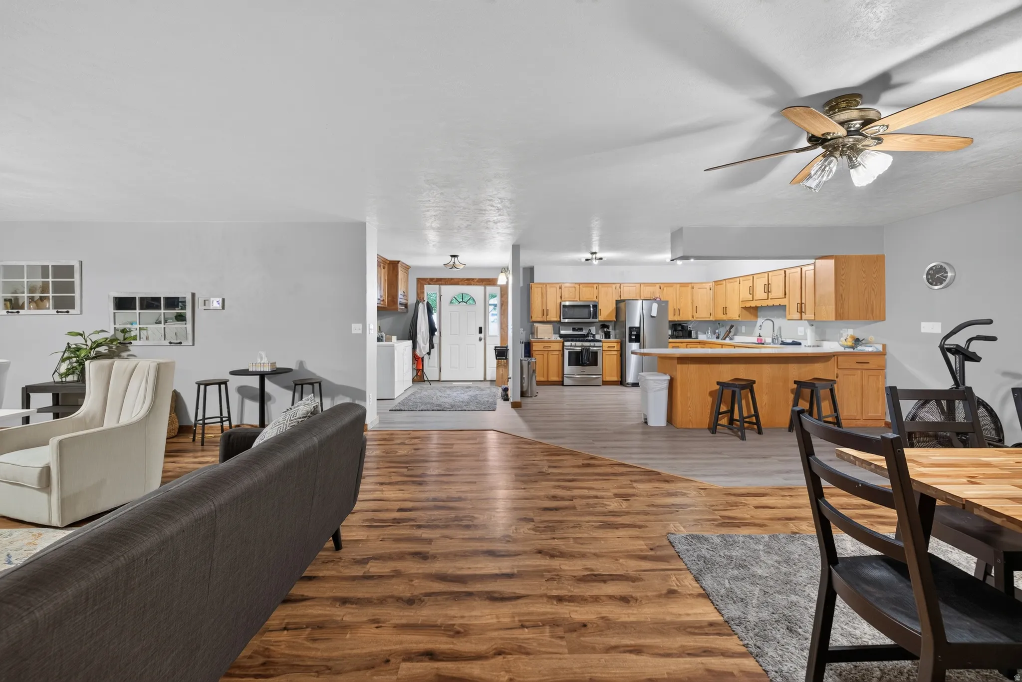 Open-concept living area featuring wood-finish flooring, a contemporary ceiling fan, and a kitchen with stainless steel appliances, light wood cabinetry, and a breakfast bar