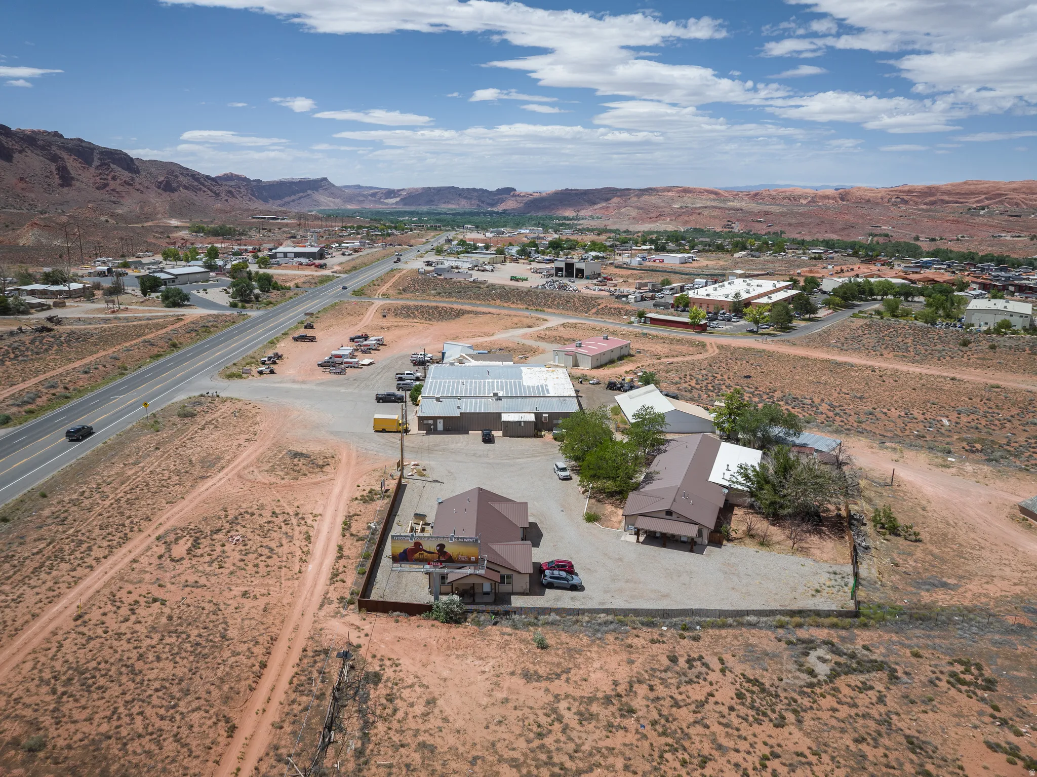 Commercial property featuring multiple buildings with brown roofs, a large gravel parking area, and a fenced perimeter