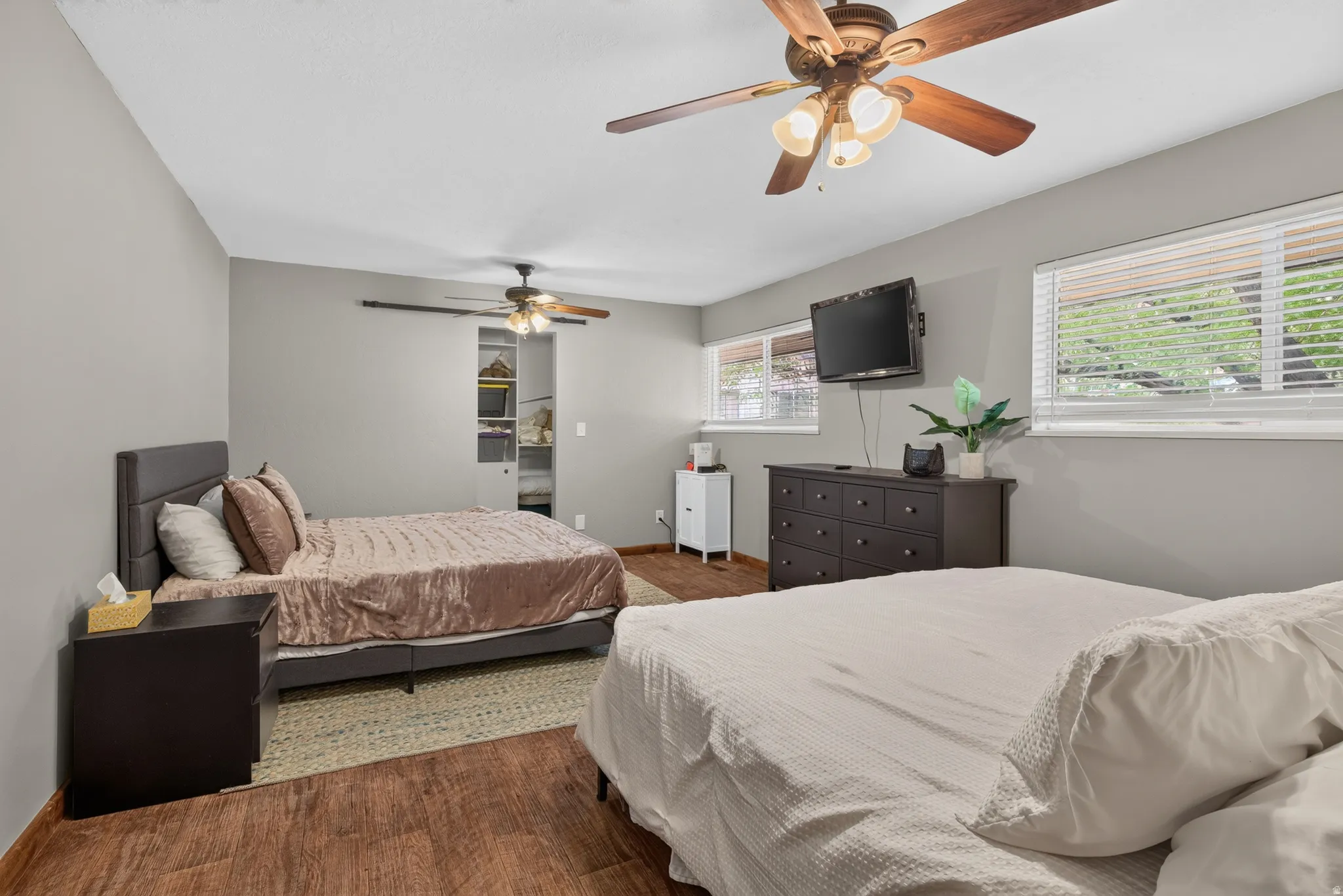Spacious bedroom featuring wood-finish flooring, two large windows with blinds, and two ceiling fans with integrated lighting