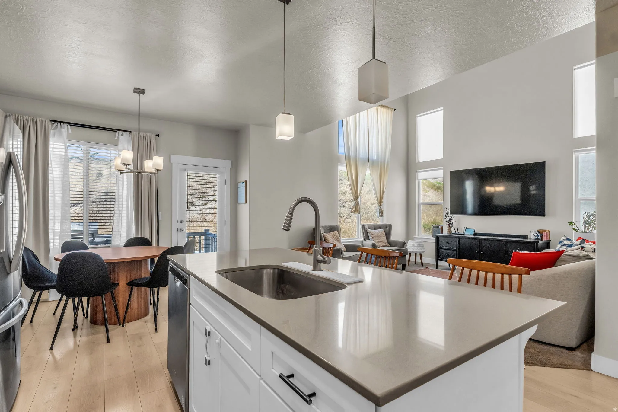 Spacious open-concept living area featuring a kitchen island with sink, light wood-finish flooring, high ceilings with pendant lighting, and multiple large windows