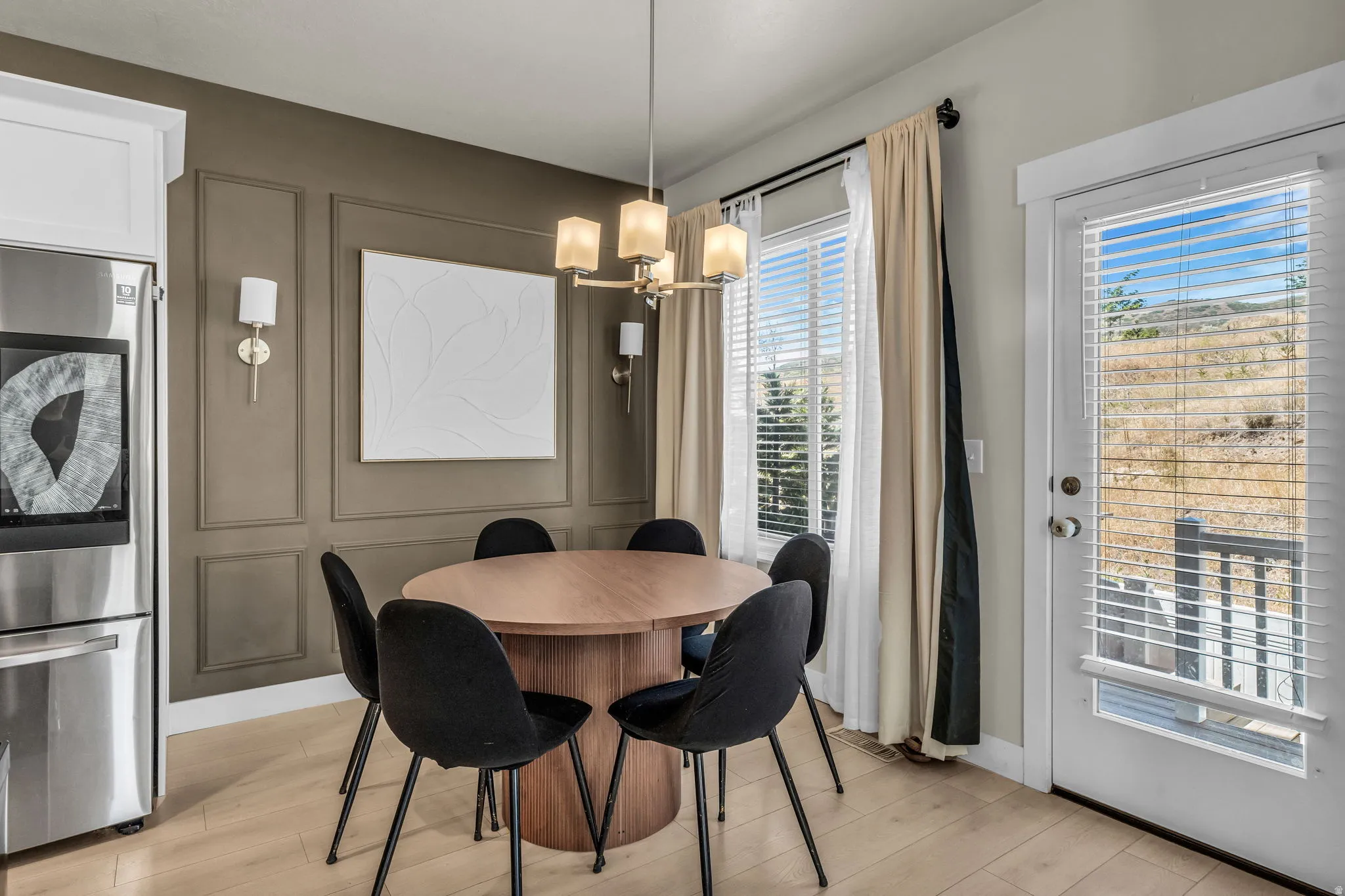 Dining area featuring wood-finish flooring, a contemporary chandelier, two windows with blinds, a white door with blinds, and a built-in stainless steel refrigerator