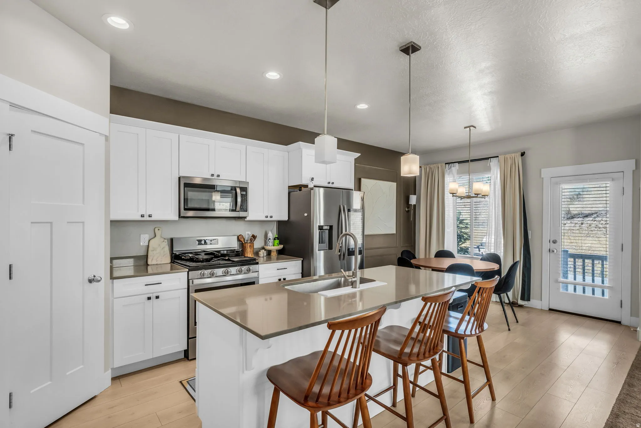 Kitchen featuring an island with seating, stainless steel appliances, white cabinetry, light wood-finish flooring, and a dining area with a glass-paned door opening to a deck