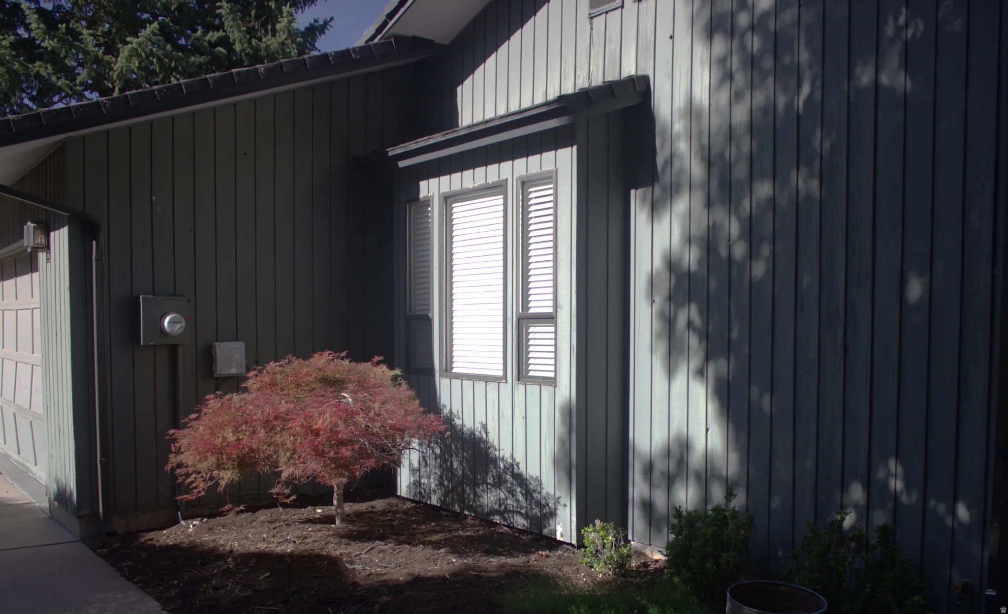 Exterior facade featuring vertical siding, bay window with louvered blinds, attached garage, and mature landscaping