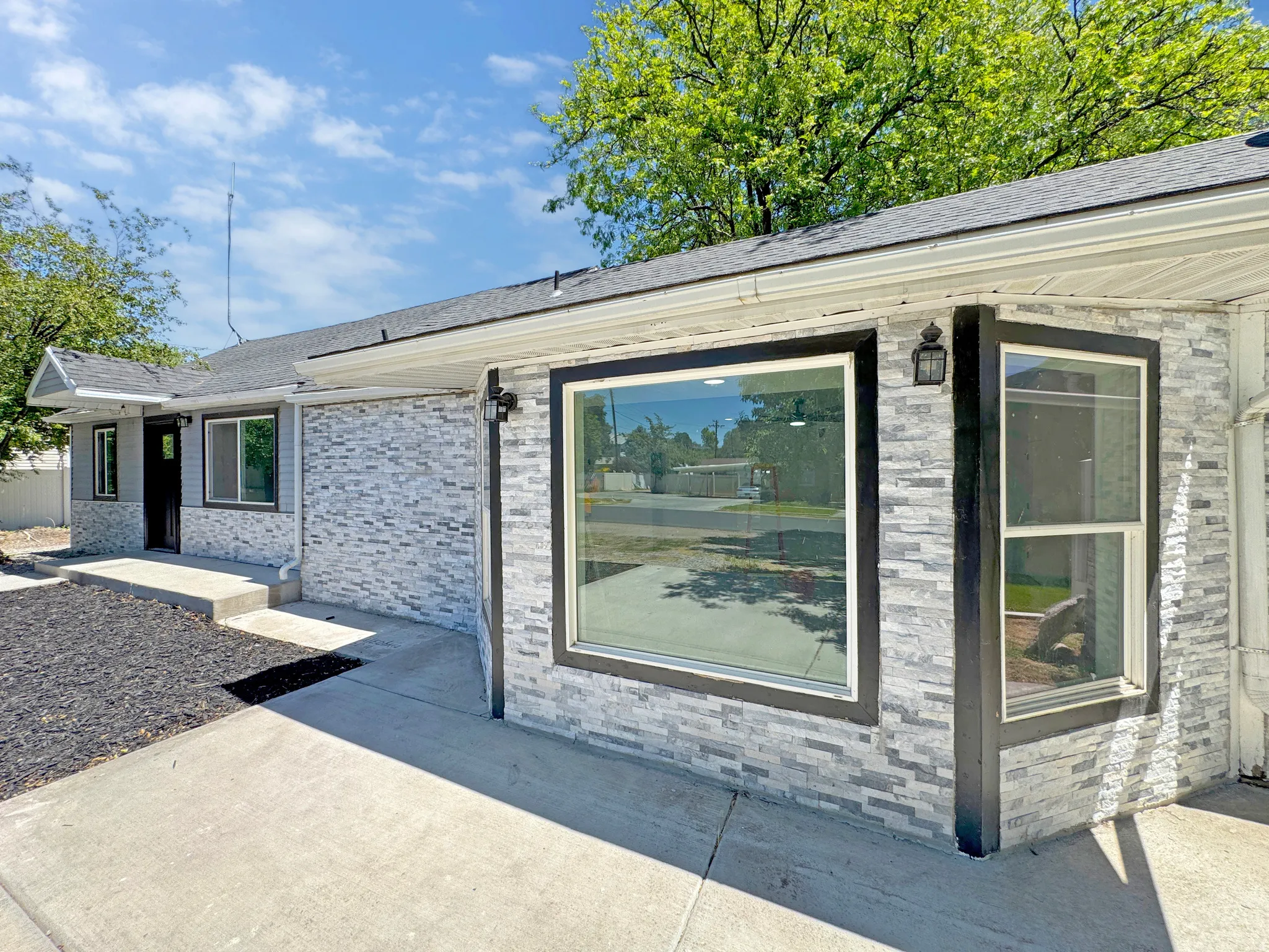 Exterior featuring grey stacked stone veneer, a dark shingle roof, dark window trim, and exterior sconce lighting