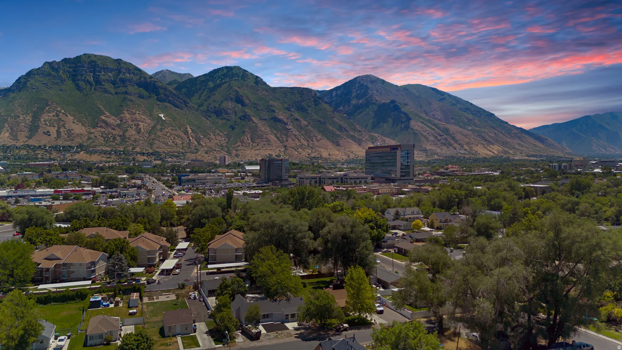 Expansive aerial perspective showcasing a mountainous backdrop, a verdant urban landscape with mature trees, a mix of residential structures, and commercial buildings