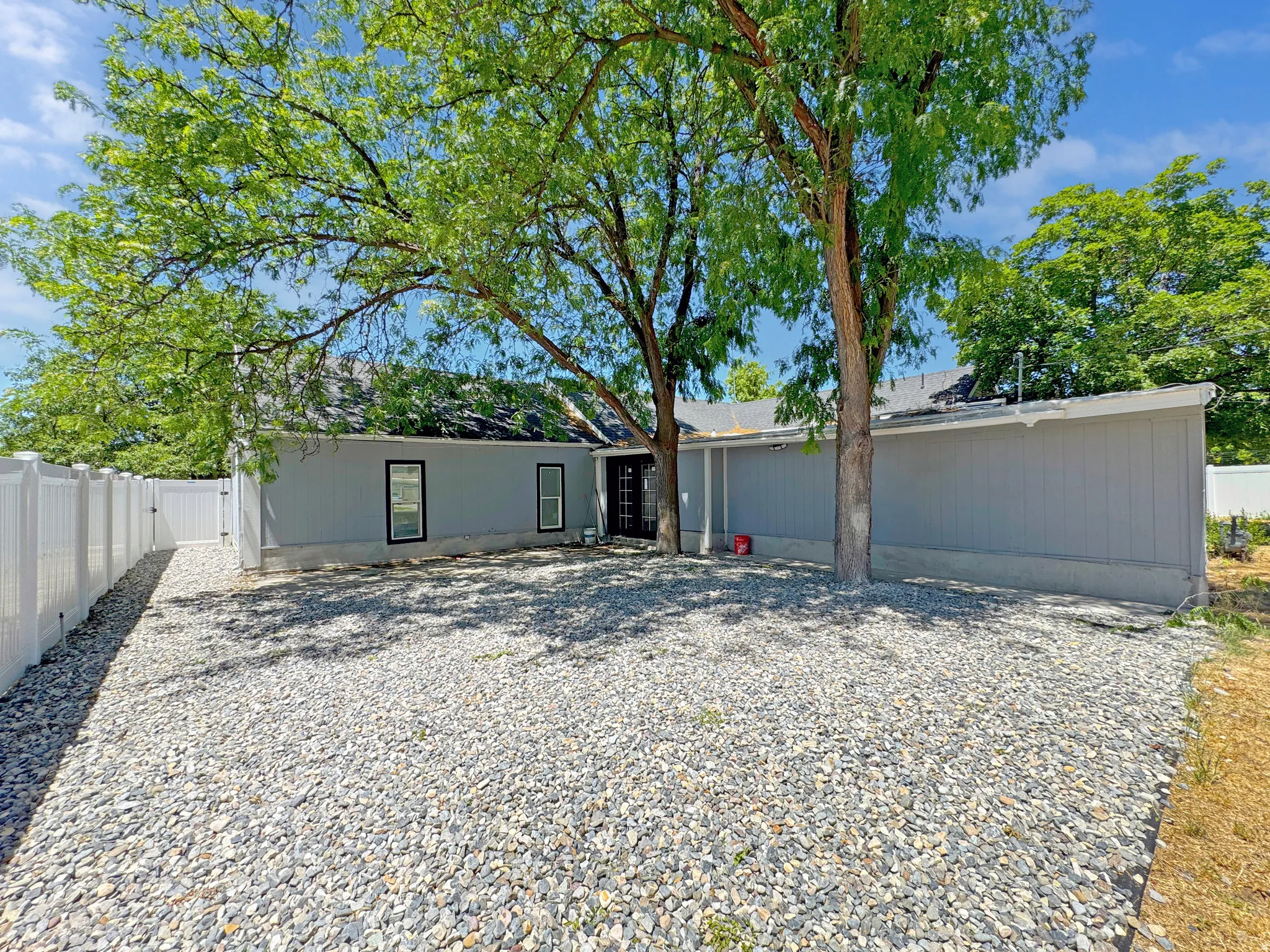 Rear exterior featuring light gray siding, a dark roof, and multiple mature trees providing natural shading