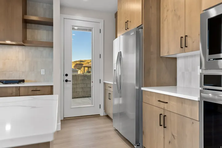 Modern kitchen featuring light wood-finish cabinetry with black hardware, stainless steel appliances, white countertops, and a full-light door providing access to exterior space