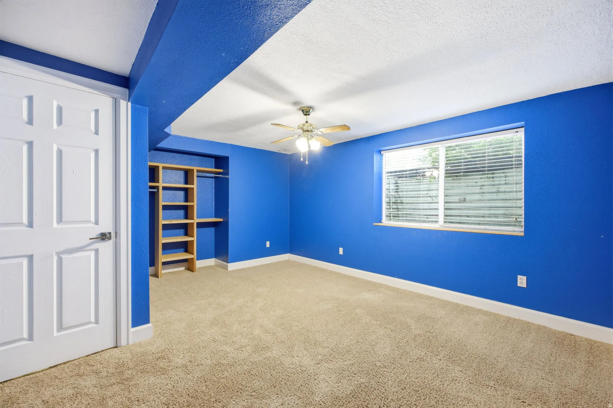 Carpeted room featuring a built-in closet system with shelving and hanging rods, a window with horizontal blinds, a ceiling fan with integrated lighting, and a white six-panel door