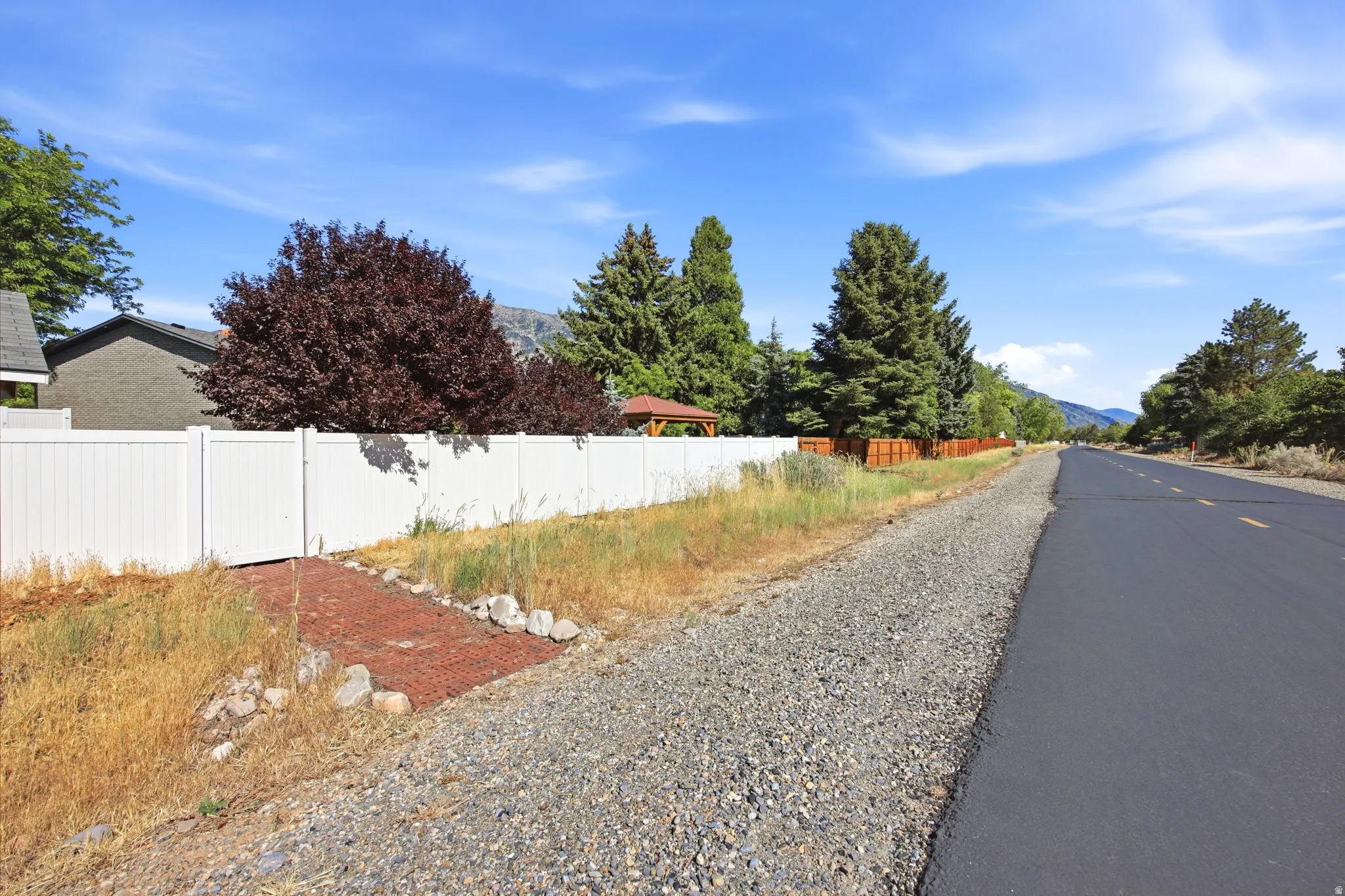 White vinyl privacy fence alongside a paved road, featuring a red gravel pathway entrance