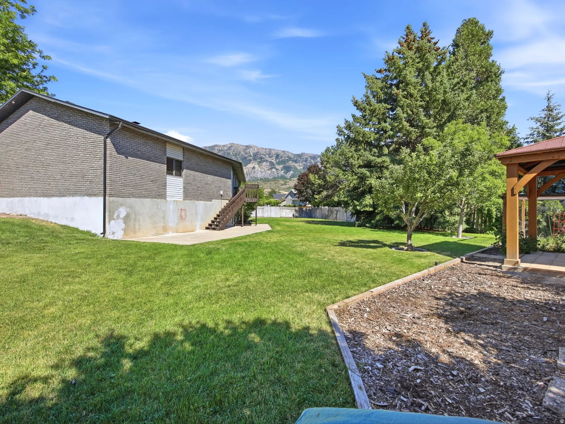 Expansive grassy backyard featuring a built-in gazebo with a red roof, mature evergreen trees, and a wooden staircase leading to the upper level of the property