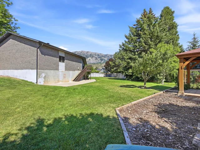 Expansive grassy backyard featuring a built-in gazebo with a red roof, mature evergreen trees, and a wooden staircase leading to the upper level of the property