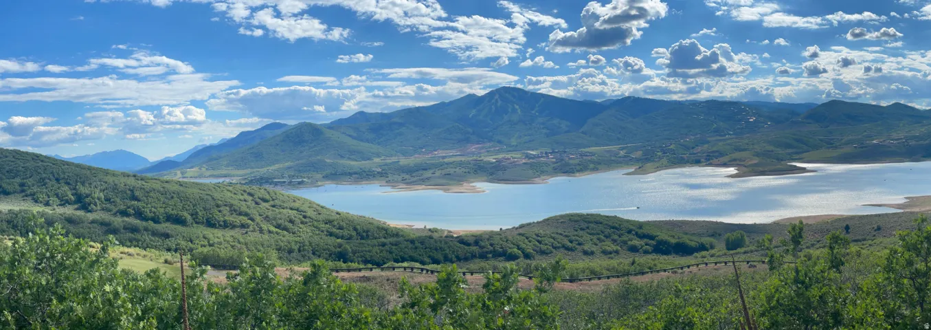 Expansive mountain panorama featuring a significant body of water, rolling green hills, and a clear sky with scattered clouds