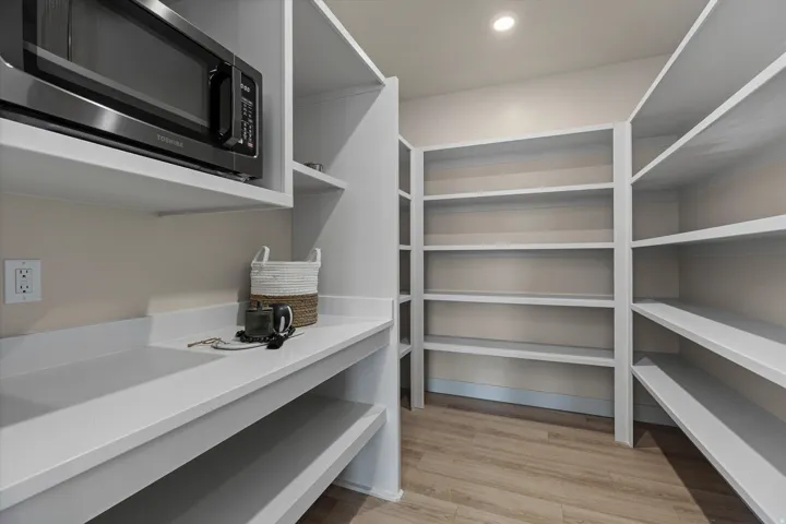 Thoughtfully designed pantry featuring extensive shelving, a built-in microwave, white countertops, wood-finish flooring, and recessed lighting