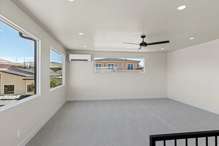 Spacious room featuring light gray carpeting, recessed ceiling lighting, a contemporary ceiling fan, multiple windows, and a wall-mounted climate control unit