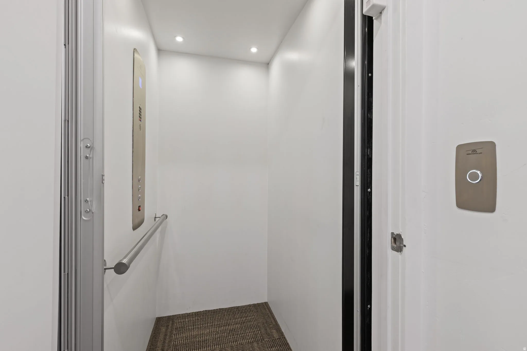 Interior residential elevator featuring recessed lighting, a brushed metal control panel, a stainless steel handrail, and woven carpet flooring