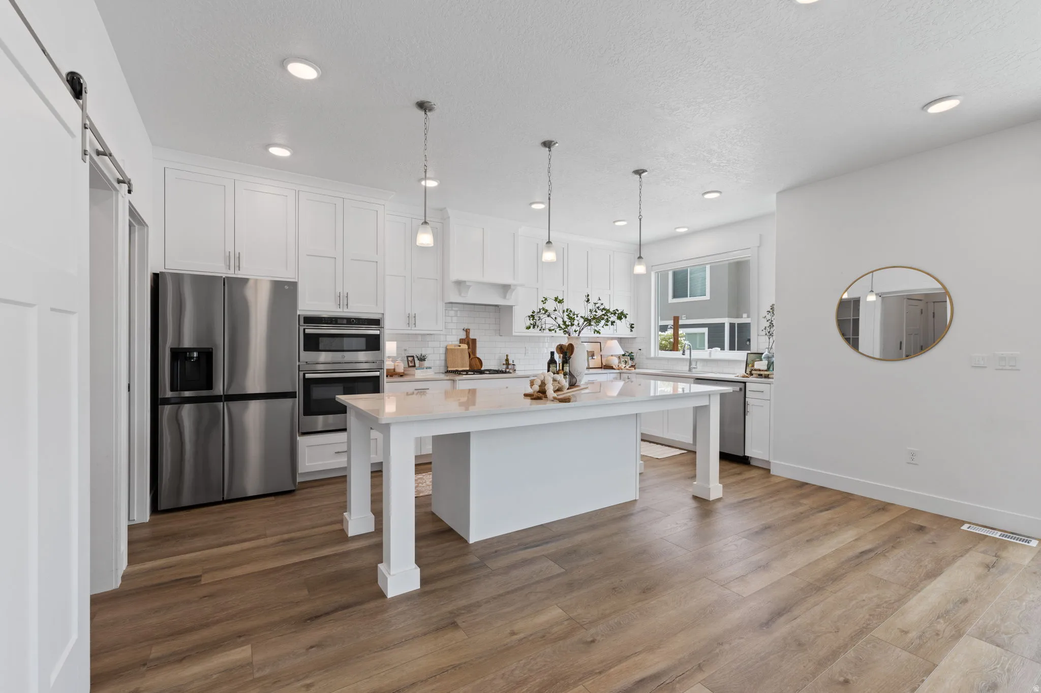 Gourmet kitchen featuring a large central island, white shaker cabinetry, stainless steel appliances including a double oven and refrigerator, wood-finish flooring, and recessed lighting