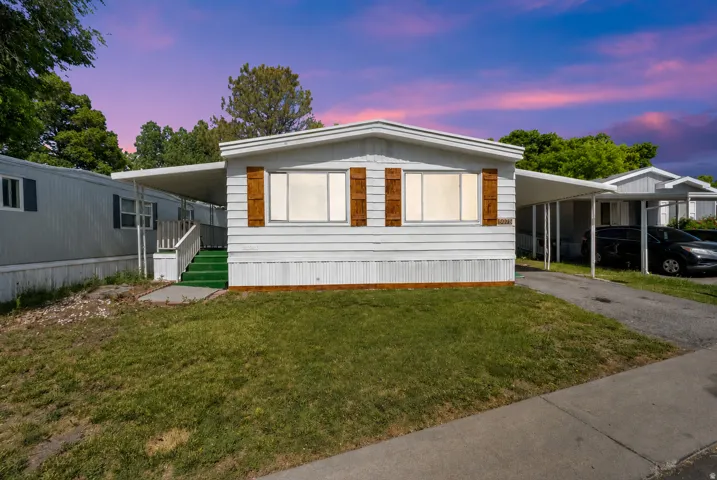 White manufactured home featuring horizontal siding, brown decorative shutters, a covered entry with green steps, a front lawn, and an attached carport