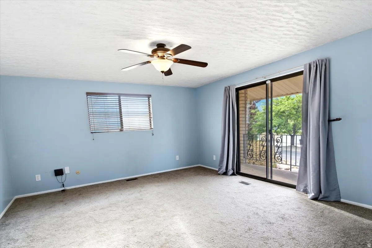 Light blue painted walls, ceiling fan with integrated lighting, window with horizontal blinds, sliding glass door, and a wrought iron balcony railing