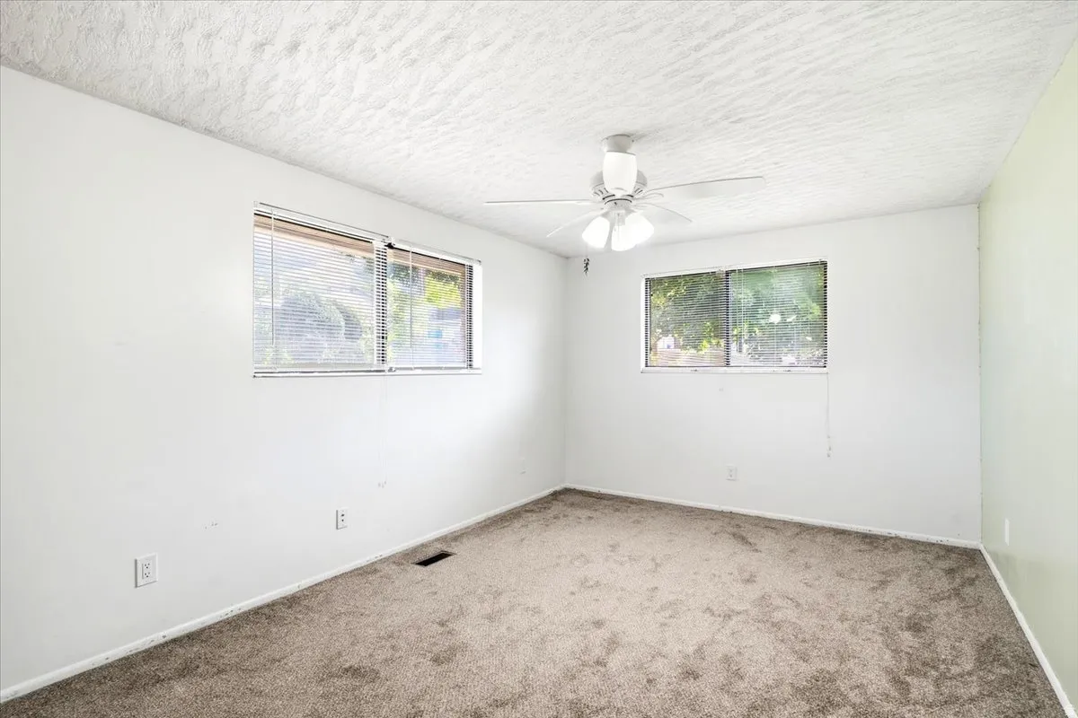 Neutral-toned carpeted flooring, white painted walls, textured ceiling, a ceiling fan, and two windows with blinds