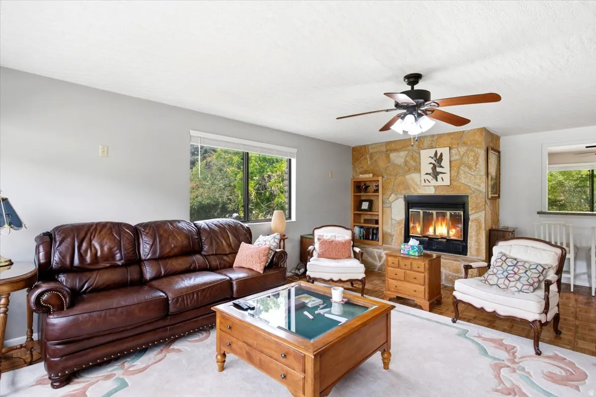 Living area featuring a stone-clad fireplace with a black metal insert, a ceiling fan with integrated lighting, and a large window offering exterior views