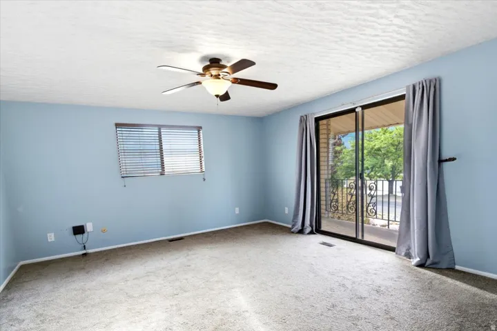 Light blue painted walls, ceiling fan with integrated lighting, window with horizontal blinds, sliding glass door, and a wrought iron balcony railing