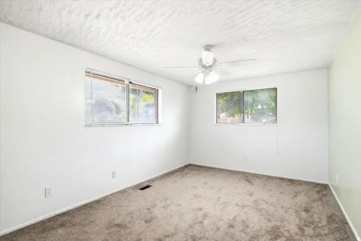 Neutral-toned carpeted flooring, white painted walls, textured ceiling, a ceiling fan, and two windows with blinds