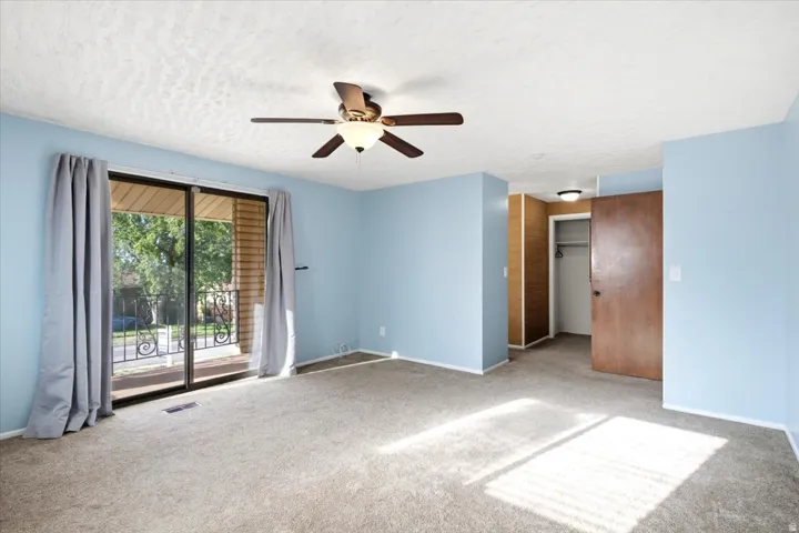 Sliding glass doors with a decorative metal balcony railing, ceiling fan with integrated lighting, wood-finish interior door, and carpeted flooring