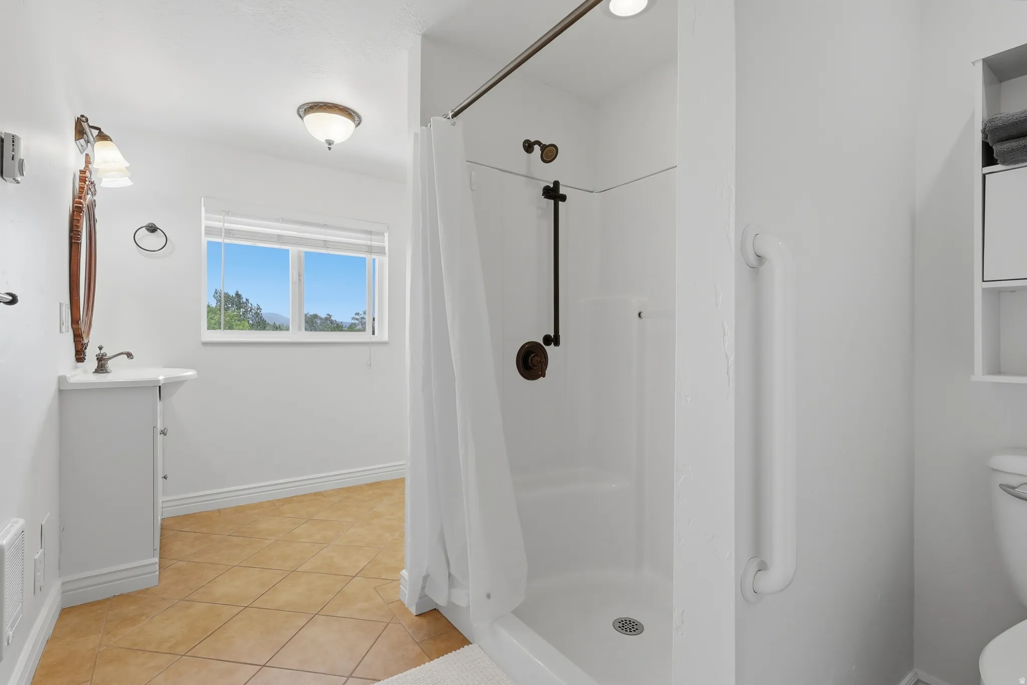 Bathroom featuring a stand-up shower with a bronze fixture, a white vanity with an oval mirror, and a window with a natural backdrop