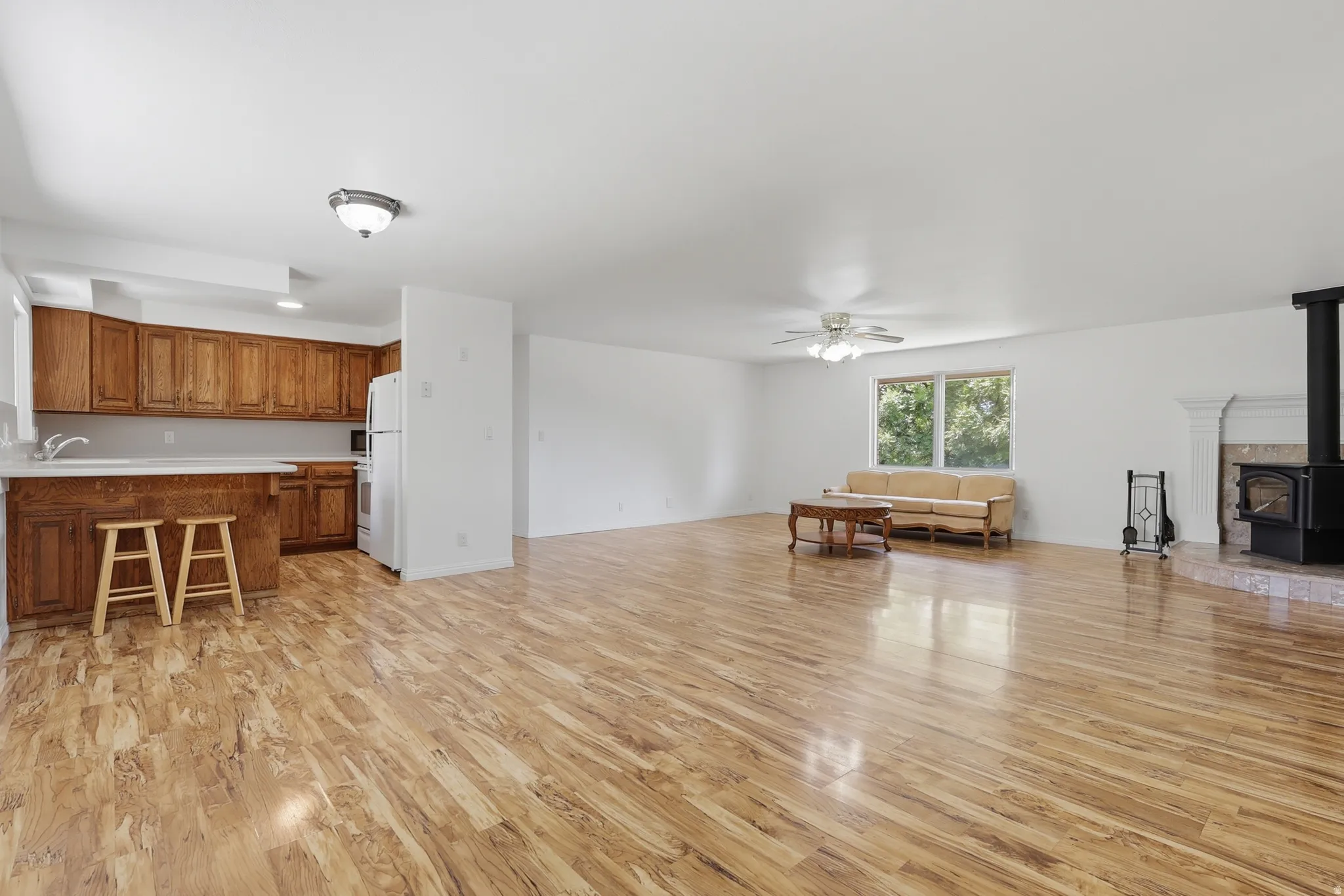 Spacious open-concept living area featuring wood-finish flooring, a white ceiling fan, and a prominent wood-burning stove with a tiled hearth and decorative mantel