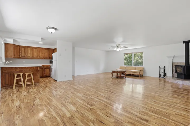 Spacious open-concept living area featuring wood-finish flooring, a white ceiling fan, and a prominent wood-burning stove with a tiled hearth and decorative mantel