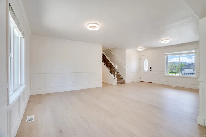 Expansive open-concept room featuring light wood-finish flooring, wainscoting paneling, recessed lighting, a front entry door with an oval glass insert, and a staircase with wrought iron balusters