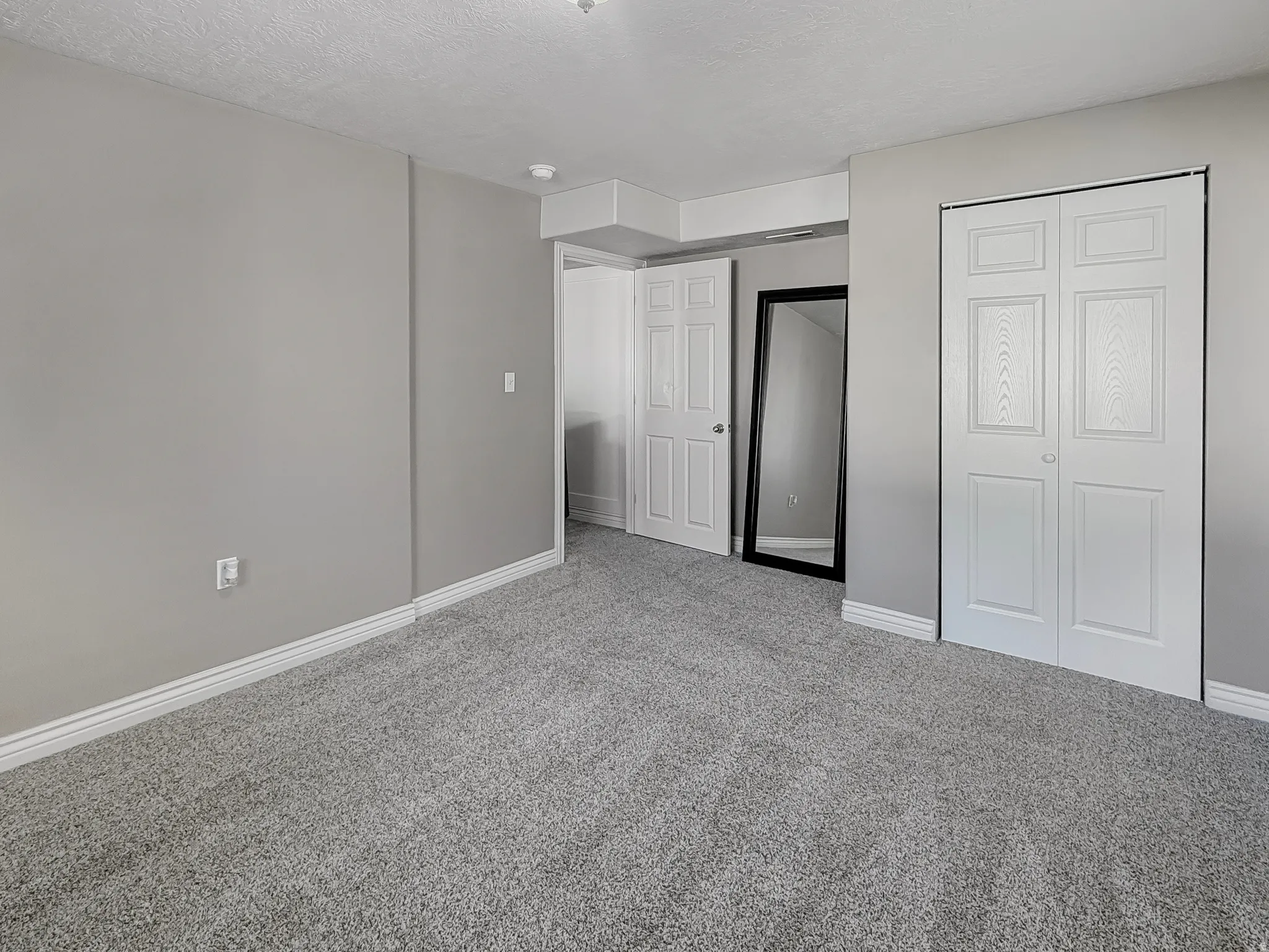 Spacious interior room featuring light gray wall paint, gray carpeting, white baseboards, and white bi-fold closet doors